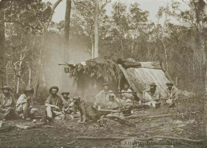 Eight men and a dog around a campfire outside a rudimentary shelter in north Queensland in the 1860s